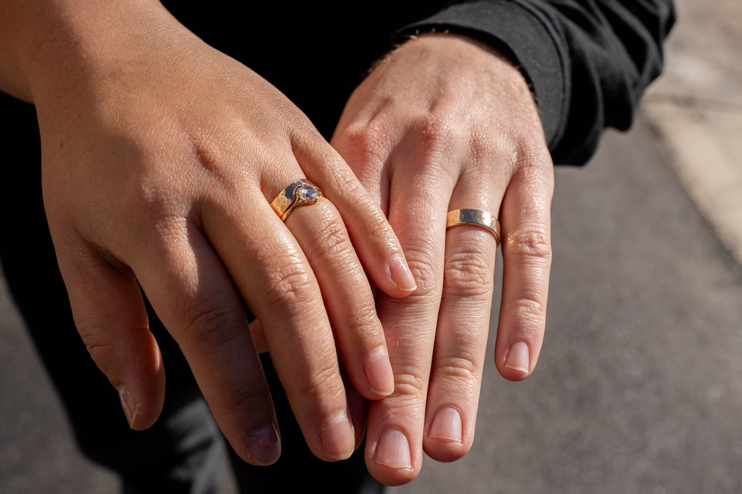 Custom shadow band in hammered gold wrapping around an unconventional engagement ring, photographed on joined hands in daylight.