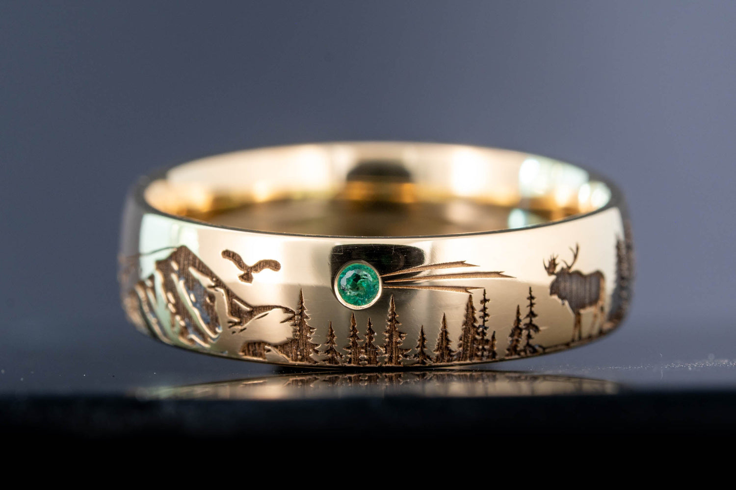 Laser engraved 18k yellow gold ring featuring a mountain landscape, wildlife silhouettes, and a flush set emerald meteor detail.