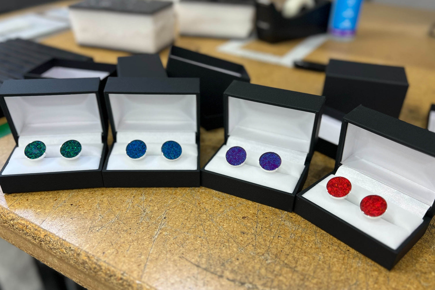 Four sets of custom sterling silver opal cufflinks in black gift boxes on the Honest Hands workshop bench, each set in a different opal color for the groom and groomsmen