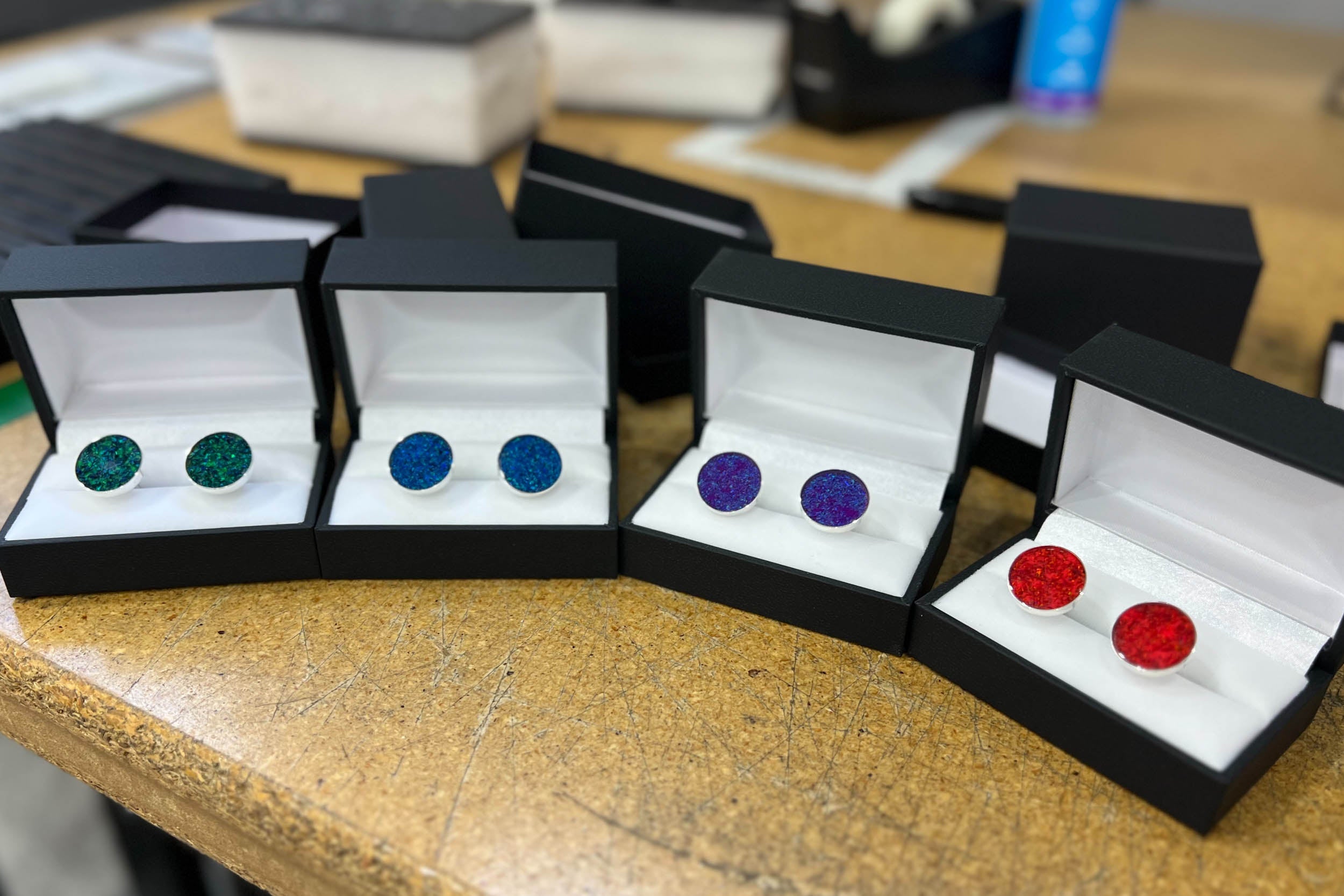Four sets of custom sterling silver opal cufflinks in black gift boxes on the Honest Hands workshop bench, each set in a different opal color for the groom and groomsmen